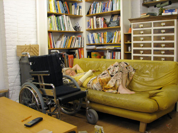 Sylvia lying on the couch, with wheelchair in front, and book cases in the back