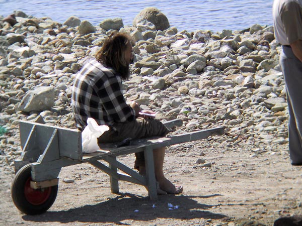 Man met baard en blote voeten zit samen met zijn plastic zak op kruiwagen