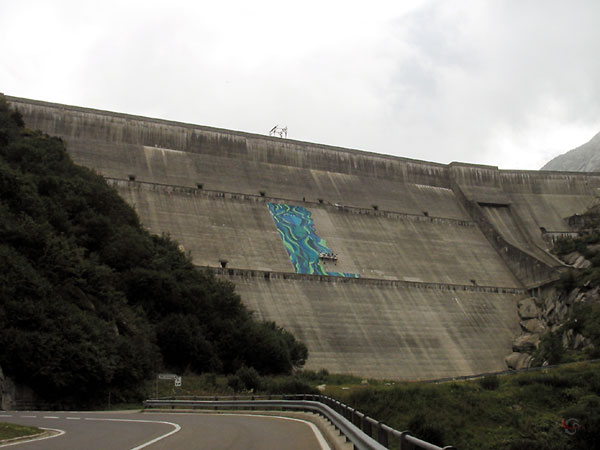 Enorme betonnen dam, van onderaf gezien; mensen in een stellage aan het schilderen