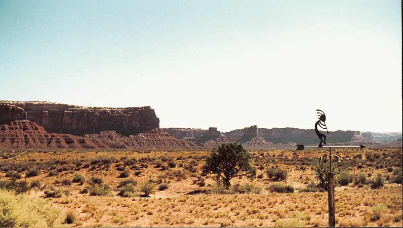 Vlakte met loodrechte rode canyonwanden, en het fluitspelende figuurtje Kokopelli op de voorgrond