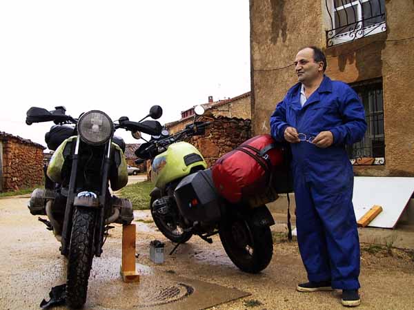 Man in blauwe overall naast de twee motoren, leemkleurig huis op de achtergrond