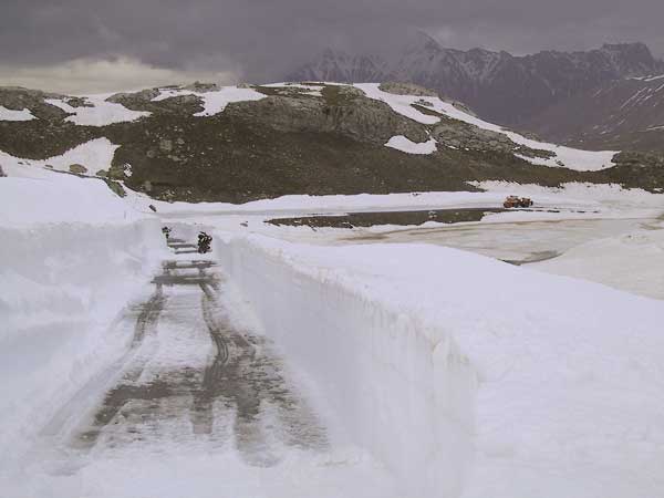 Besneeuwde weg tussen sneeuwwanden, besneeuwde bergen, geparkeerde motor in de verte